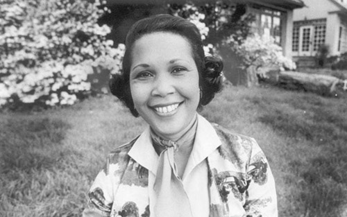Black and white photo of Barbara Johns smiling in front of flowering bushes.